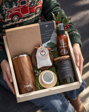 A wooden gift box held by a person wearing a green Christmas sweater with a red truck pattern. The box contains a wood-grain insulated tumbler, a candle, a small woven bag, a brown bottle, and a black jar, arranged on green moss.
