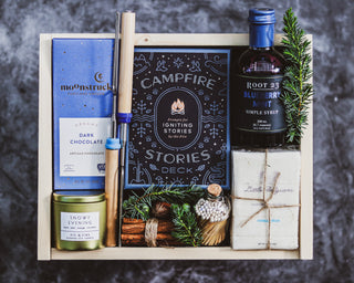 A gift box containing handcrafted vanilla marshmallows, wooden roasting sticks, a chocolate bar, a soy candle, blueberry syrup, matches in a glass jar, and a deck of storytelling prompts.