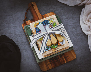 Charcuterie recipe cards tied with a ribbon on a wooden cutting board and towel; finished with cheese tool set