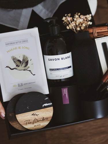 Luxury collection of kitchen items including dish soap, custom engraved coasters, vase, candle and wooden tools in a dark matte gift box.