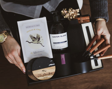 Luxury collection of kitchen items including dish soap, custom engraved coasters, vase, candle and wooden tools in a dark matte gift box.