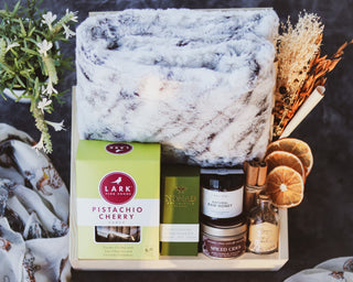 Curated gift box containing a faux fur marble throw blanket, honey, soy candle, hand sanitizer, artisan shortbread cookies, matches in a glass jar, floral bundle, potpourri, and cinnamon sticks, all arranged in a wooden basket.