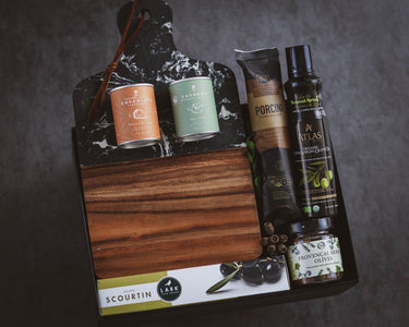 Gourmet gift set with various items including a can, bottles, and a jar on a wooden board against a dark background.
