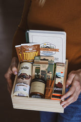 Person holding a cabin-themed gift box with camping essentials and a National Parks Journal.