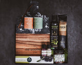 Gourmet gift set with a wooden cutting board, chocolate bars, olive oil, and other items on a dark background.