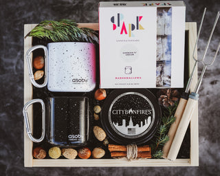 A gift box set that includes a portable fire pit, marshmallow roasting sticks, and campfire mugs, all displayed on a wooden tray.